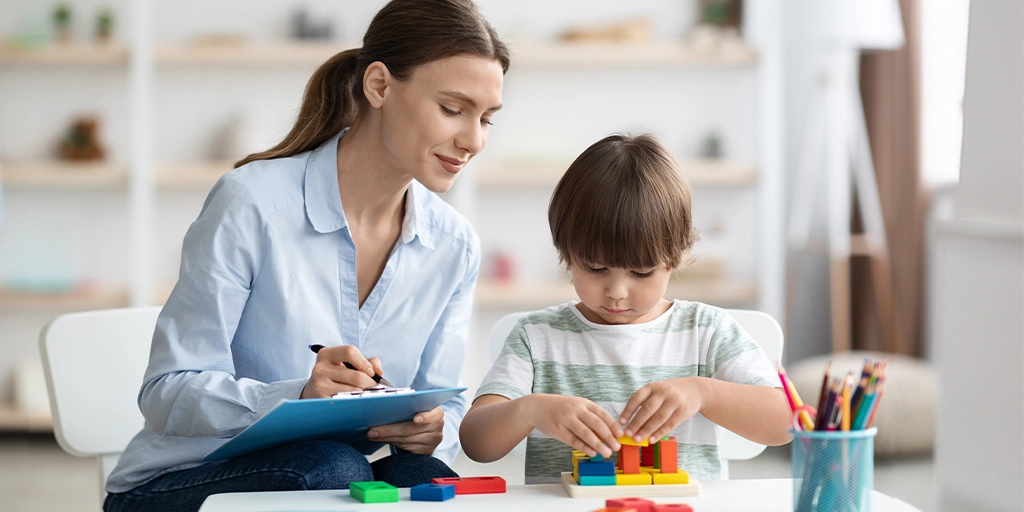 A young boy with autism focuses on building with colorful blocks while a supportive therapist or teacher takes notes beside him.
