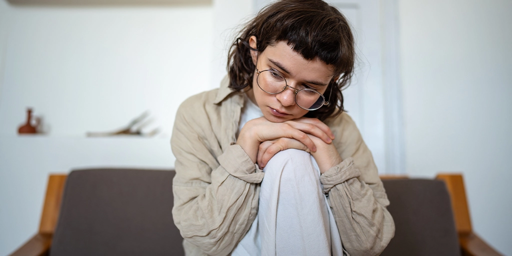A young woman sitting on a sofa, appearing thoughtful, representing the challenges of masking in autism.
