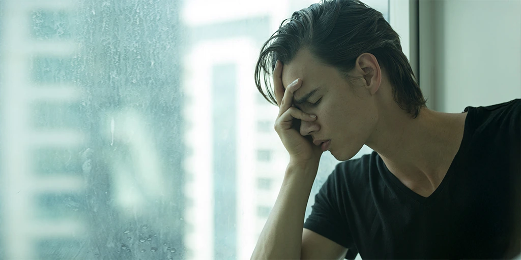 Young man sitting by a window with his hand on his face, appearing overwhelmed or anxious.<br srcset=