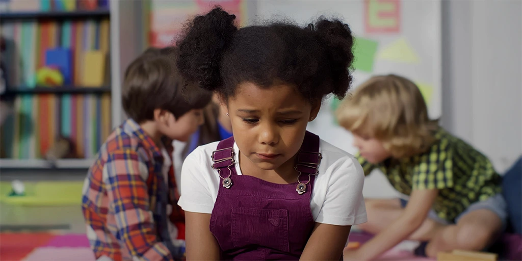 Young girl sitting alone in a classroom, looking sad while other children play in the background.